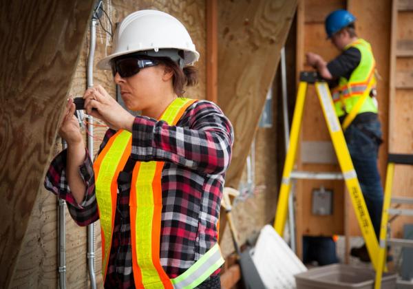 A woman wearing a hardhat, safety goggles and high visibility vest installs an electrical panel into a wall