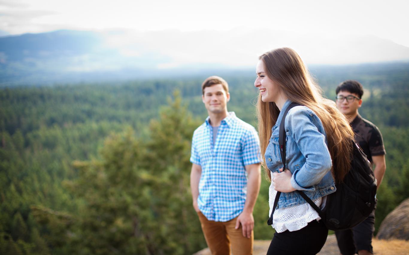 Three students stand on a cliffside looking over a vista of trees